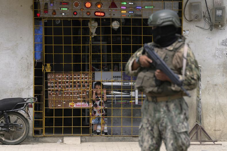 A boy watches from behind the latticework of a store as a soldier stands guard at a security post, in Durán, Ecuador, on August 14, 2023. The country's president declared a state of emergency in some areas after the murder of a presidential candidate after a rally in the campaign for the early elections, scheduled for August 20.  (AP Photo/Martín Mejía)